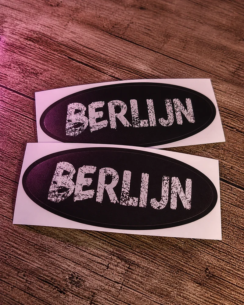 Twee zwarte ovale stickers met witte BERLIJN-opdruk op een houten ondergrond