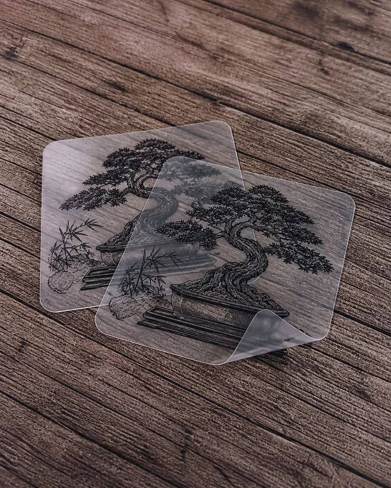 Twee transparante stickers met zwart bonsaidesign op een houten ondergrond